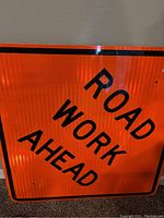 Close-up image of the orange reflective road sign with black 'ROAD WORK AHEAD' text showing texture and minor wear.
