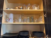 Two shelves inside a wooden cabinet with various clear drinking glasses and glass barware. The lower shelf holds black stoneware plates and bowls.