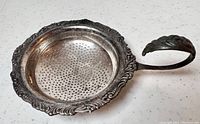 Close-up side view of tea strainer showing pierced bowl and decorative handle with embossed leaf design.