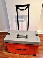 Closed toolbox suitcase showing orange body, gray lid, and extended handle with wheels.