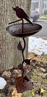Full view of cast iron bird bath outdoors on grass next to a tree, showing bird on basin and leafy stem.