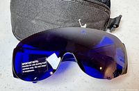 Close-up photo of blue tinted glasses from front showing wraparound lens with warning label and part of black case behind.