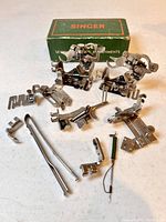Multiple Singer metal sewing machine attachments with original green box in background.