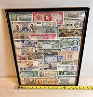 Full view of the black framed art piece containing a variety of paper money from around the world, showing arrangement and some identifiable currencies.