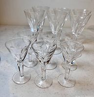 Overall view of 12 glasses including 6 smaller cordial glasses in front and 6 larger wine glasses behind on a white speckled table surface.