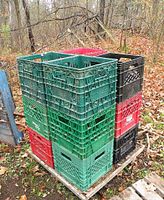 Photo of 12 plastic milk crates stacked on a wooden pallet outdoors on a dirt and leaf-covered ground. Crates are green, red, black, and teal with visible dirt and weathering.