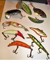 9 assorted vintage fishing lures laid out on a white surface showing various shapes, colors, and two lures having metal spinner blades.