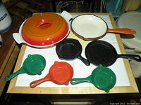 Full set of cast iron and enamel cookware items arranged on table including oven dish with lid, frying pans and child play size enamel pans.