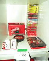 Full view of lot showing 17 decks of playing cards stacked, a red tin case for double decks, set of Coca-Cola tin coasters, and 2 miniature vending machines with cart.
