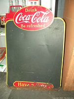 Full front view of the vintage Coca Cola tin chalk board sign showing red oval logo and black writing surface. Yellow border visible around the edges.