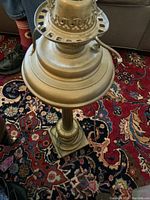 Top and side view of the antique brass oil lamp showing the wick holder and electrical cord detail, placed on a patterned carpet.