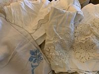 Photo showing folded white and cream linens including tablecloths and doilies with lace and embroidered details, including blue floral embroidery.