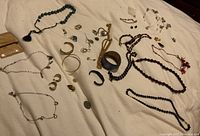 Photo showing assortment of costume jewelry on a beige fabric surface including necklaces, bracelets, pins, and earrings in gold, silver, black, and colored beads.