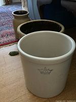 Three vintage crocks arranged on floor; large white crock in front with crown emblem, medium brown crock behind it, and smaller beige crock in back.