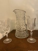 Crystal pitcher and two wine glasses shown on a wooden surface against a white wall. The pitcher has an etched floral and geometric pattern with a scalloped rim. Two matching wine glasses have faceted design.
