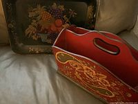 Photo showing vintage hand painted magazine holder with floral design in front and serving tray with fruit design in background on a grey surface.