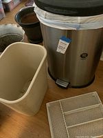 Photo showing stainless steel step-on trash can with black lid, white plastic inner liner, and white metal wire drawer organizer placed on wooden floor.