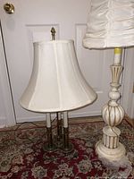 Brass table lamp with beige fabric shade and painted wood table lamp with cream fabric shade photographed together on patterned rug with white door background.