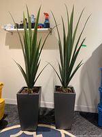 Two faux plants in tall gray planters side by side on rug against white wall