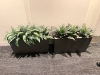 Two dark rectangular plastic planters with two different styles of faux plants, set on carpeted floor against a wall.