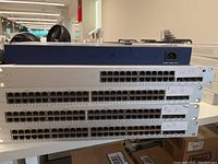 Stack of one blue Netgear GS724T and multiple silver Cisco Meraki switches labelled on rack ears