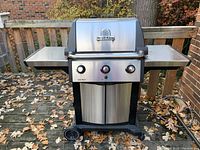 Front view of Broil King gas grill with lid closed, showing two side shelves, wheels, and double-door storage cabinet below.
