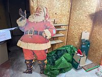 Photo of vintage Santa standee in basement setting beside green bag containing Christmas tree and two small fibre optic trees in boxes.