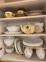 Cabinet shelf with stacked Corelle dishes, bowls, cups, saucers, and ceramic teapot with tree and script design