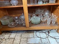 Shelf showing stacked clear glass dinner and salad plates, several glass bowls and a set of etched cornflower stemmed glasses.