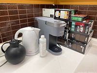 Keurig coffee maker, white electric kettle, black insulated coffee carafe, bottle of de-scaling liquid, and McCafé coffee pods on kitchen counter