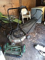 Photo showing Briggs and Stratton gas lawn mower with attached grass catcher bag and one manual reel push mower stored in a wooden shed on dirt floor.