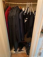 Menswear items hanging on hangers inside a small closet. Includes suit jackets in dark colors and blue, a red garment bag, and other hanging items.