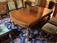 Wooden dining table with polished brown finish, ornate turned legs, surrounded by upholstered chairs, showing surface wear