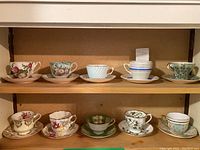 Overview of 10 vintage teacups and matching saucers arranged on two shelves showing variety of floral and striped designs