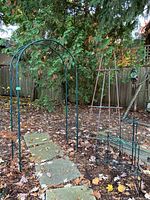 Outdoor view of the green metal garden arch trellis standing over stone pavers with dried leaves scattered on the ground, surrounded by trees and a wooden fence.