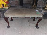Full view of the bench showing its beige upholstered seat and dark carved wooden legs with claw feet on a stone background.