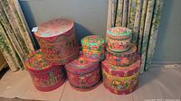 Photo showing seven colorful round and one hexagonal hand-painted boxes with folk art designs stacked and grouped together, placed on a cloth surface near curtains.