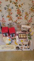 Full overview of lot items arranged on floor including duck cookie jar, boxed stir fry wok, wine stoppers, candles, magnets, lantern decorations and place card holder.