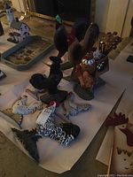 Nine varied decorative chicken figurines arranged on a table posed on white paper; includes large multicolored roosters and hens of metal and resin.