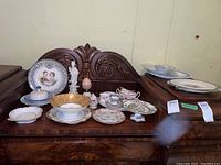Wide shot showing entire assortment of china on wooden furniture, includes various plates, bowls, figurines, and covered dish.