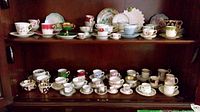 Two wood cabinet shelves displaying assorted fine china cups, saucers, plates, and a sugar bowl