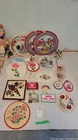 Photo showing overall collection of various embroidery hoops with farm animal scenes, floral cross stitch, small embroidered patches and quilted items arranged on a flat surface.