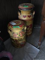 Two large decorative metal milk cans painted with flowers and hearts on a yellow background, some dents visible, set on floor.