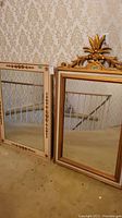 Two framed rectangular mirrors leaning against a wall with patterned wallpaper. One mirror has an ornate gold crest; the other has a cream frame with small gold accents.