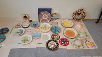 Full overview of 20 various decorative and novelty plates laid out on a white sheet showing different sizes and diverse designs including floral, animals, and holiday themes.