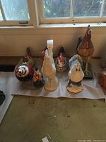 Eight varied ceramic rooster and chicken statues arranged on the floor by a window, showing different sizes and colorations including white, speckled, and colorful roosters.