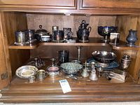 Wide shot of all silver plate items arranged on shelves inside a wooden cabinet, showing variety including bowls, candle holders, and small containers.
