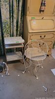 Photo showing the white metal chair with curved designs and the 3-tiered wooden table with a marble top, placed near a cabinet and curtains.