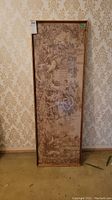 Full view of the framed tapestry stood upright against wallpapered background.