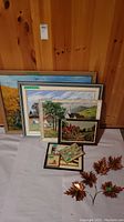 Photo showing all eight pieces of wall art including framed paintings, framed needlepoint/cross-stitch art, collage framed art, and metal maple leaf decor arranged on a white sheet against wooden wall.